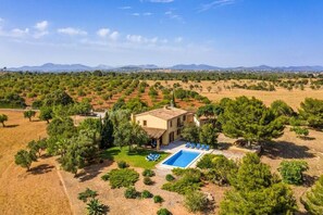 A villa with a swimming pool, surrounded by olive groves and trees, with mountains in the background.