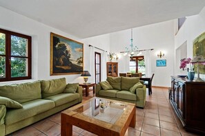A living room with two green sofas, a wooden coffee table, a painting on the wall, and a dining area in the background.