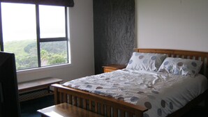 A bedroom with a large window, a bed with patterned bedding, a wooden headboard, a bedside table, and a bench.