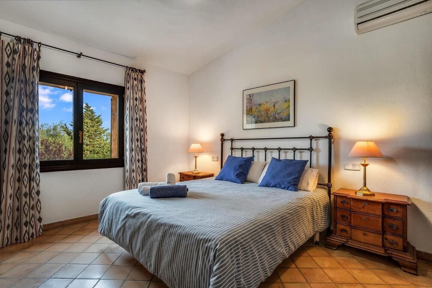 Een slaapkamer met een bed, twee nachtkastjes, een raam met gordijnen, een schilderij aan de muur en een betegelde vloer.