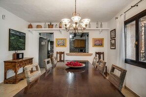 Un comedor con una mesa de madera, sillas de mimbre, una araña y armarios de cocina al fondo.