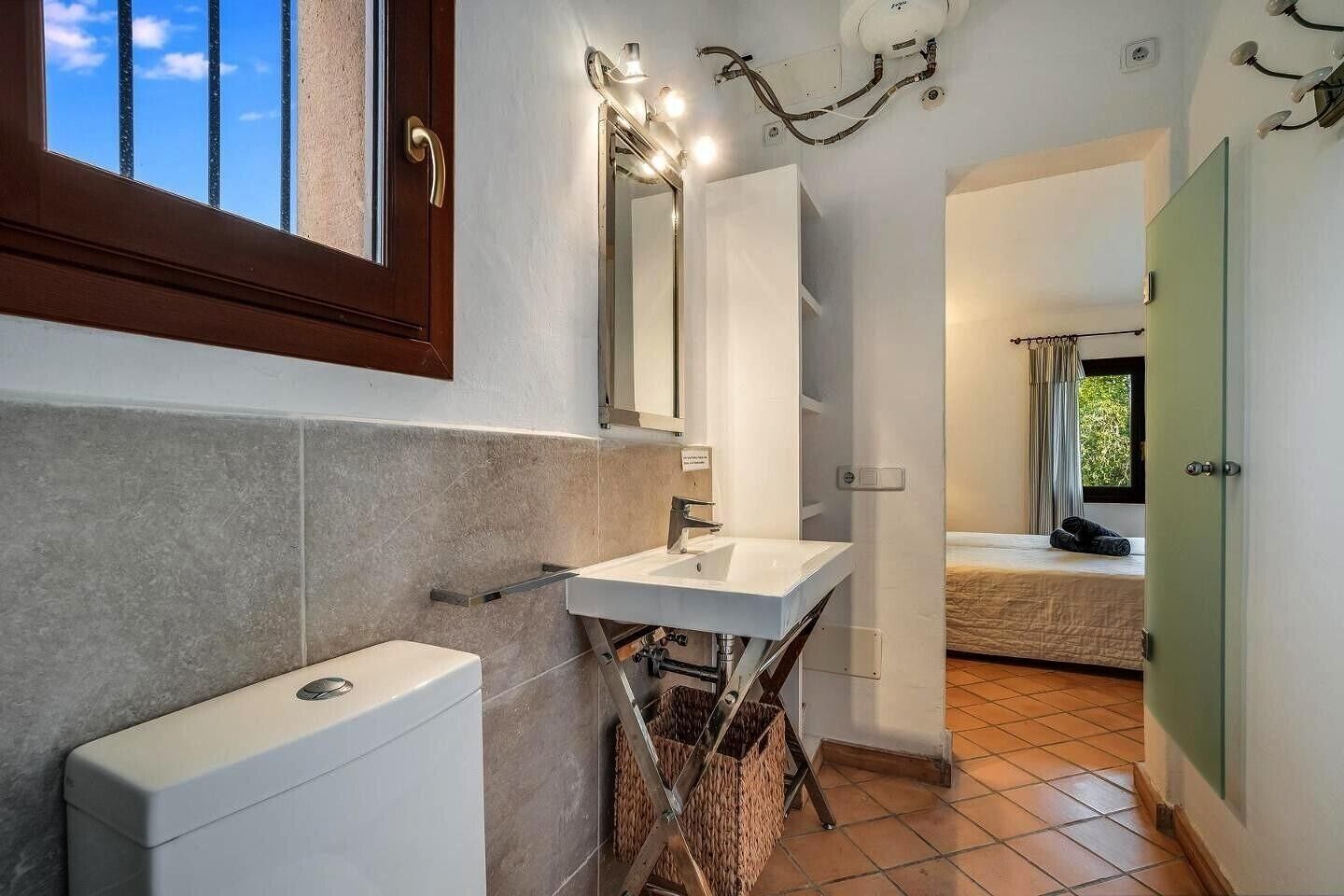 Un bagno con un water, un lavandino e uno specchio. C'è una finestra con tende e un letto nella stanza adiacente.