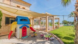 Zona de juegos infantiles con tobogán y estructura para escalar, un área de descanso cubierta y una piscina al fondo.