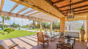 Un patio al aire libre con mobiliario de madera, una mesa de vidrio y vistas a una piscina y vegetación.