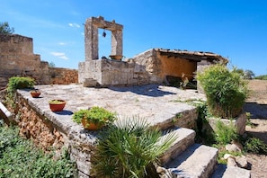 Une ancienne structure en pierre, dotée d’une petite porte en arc de cercle, de pots en terre cuite ornés de plantes et d’un bâtiment rustique surmonté d’un toit en tuiles.