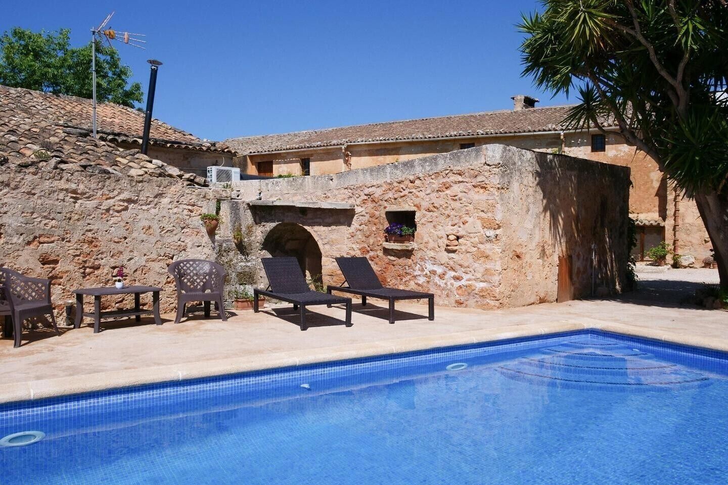 Una piscina con sillas de descanso y una mesa, un muro de piedra y un cielo azul despejado.