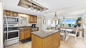 A kitchen with granite countertops, wooden cabinets, a large island, and a dining area with a view. - Back Bay 1106 | Panoramic Views | Spacious Waterfront Penthouse | My Beach Getaways (Orange Beach)