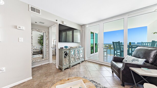 A spacious living area with a large TV, a leather sofa, and a balcony with a city view. - Back Bay 1106 | Panoramic Views | Spacious Waterfront Penthouse | My Beach Getaways (Orange Beach)