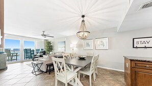 A dining area with a large table, white chairs, a chandelier, and a view of the ocean through large windows. - Back Bay 1106 | Panoramic Views | Spacious Waterfront Penthouse | My Beach Getaways (Orange Beach)