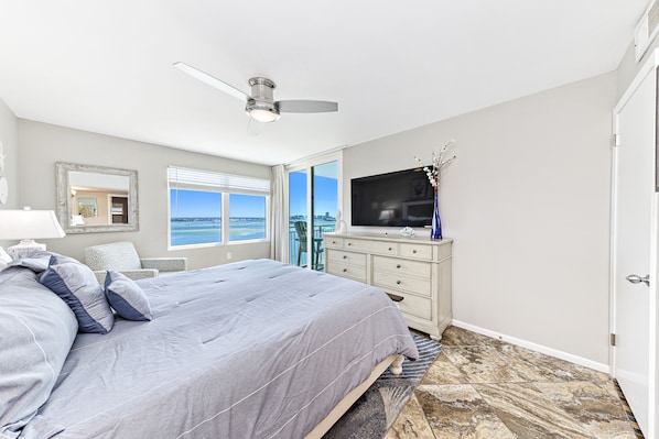 A bedroom with a bed, a TV, a dresser, a ceiling fan, and a view of the city.