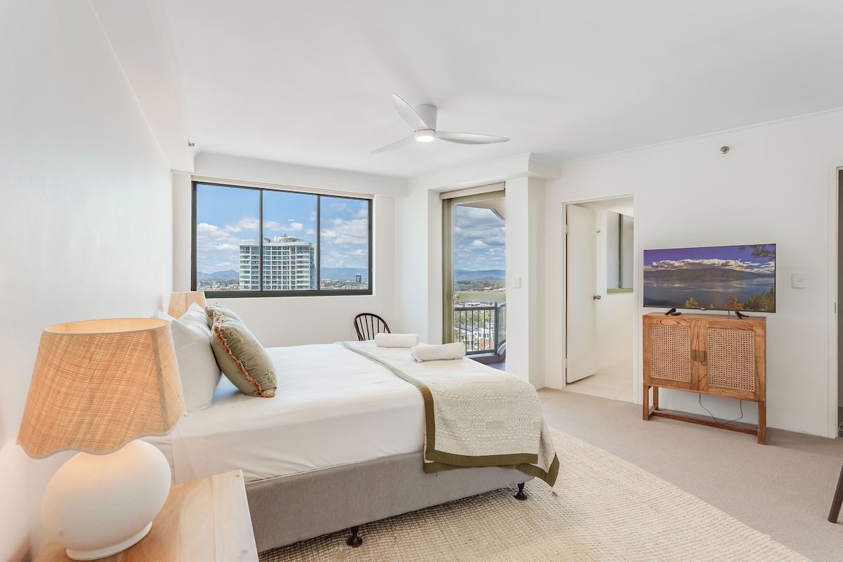 A bedroom with a bed, a ceiling fan, a TV on a wooden stand, and a large window with a city view.