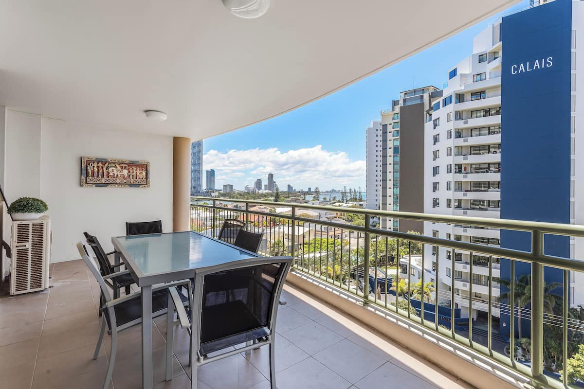 A balcony with a glass table and chairs, overlooking a cityscape with modern buildings and a clear sky.