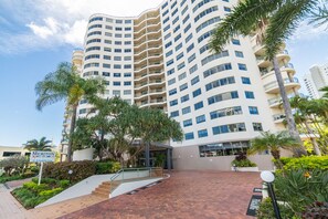 Un hotel de varios pisos con fachada curva, balcones y un área de entrada ajardinada con palmeras y arbustos.