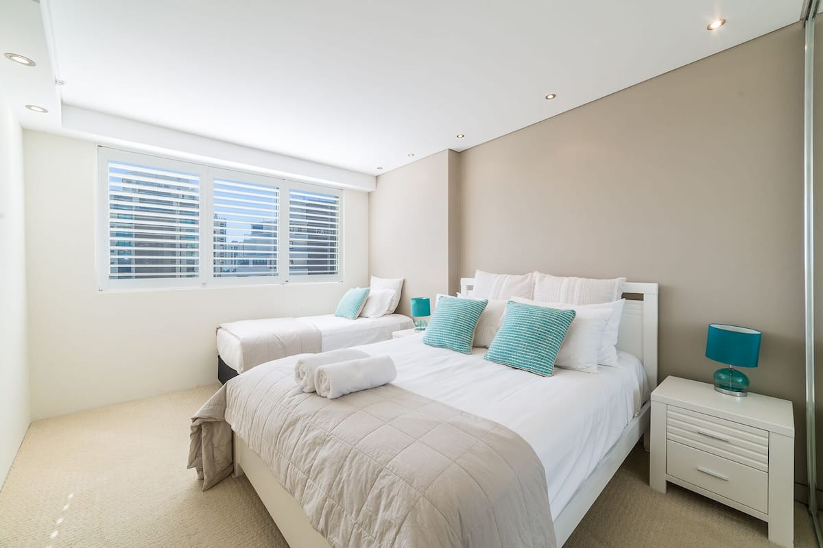 A modern bedroom with two beds, a nightstand, and a window with blinds.