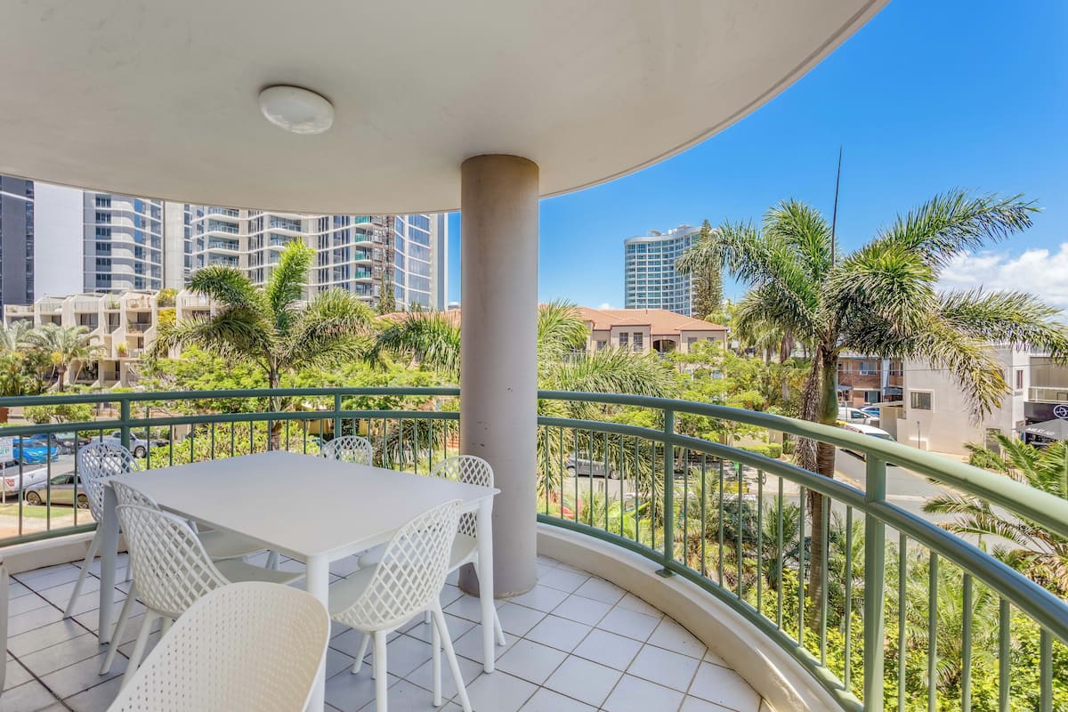 A balcony with a white table and chairs, surrounded by a green railing, overlooking a cityscape with palm trees and buildings.
