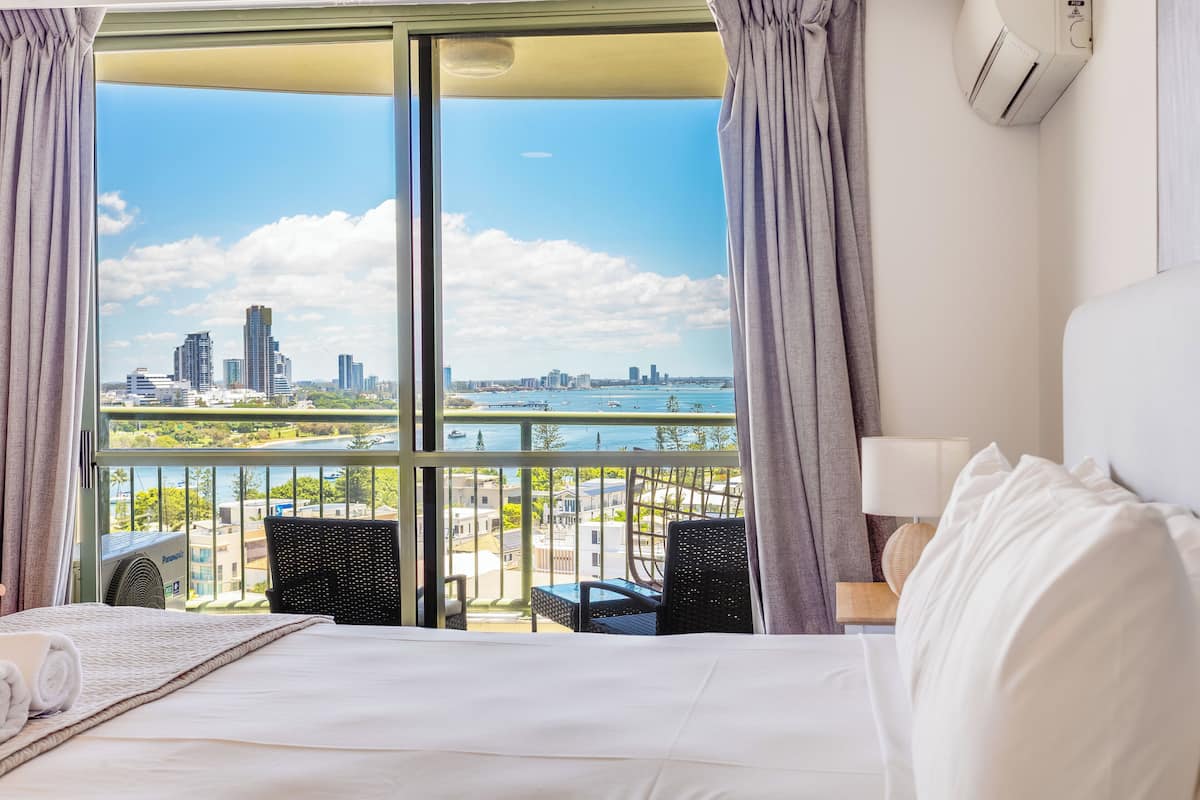 A hotel room with a bed, a view of a city skyline, and a balcony with chairs.