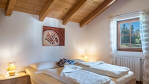 Un dormitorio con techo de madera, una cama con ropa blanca, una mesita de noche con lámpara, un cuadro en la pared y una ventana con vistas a árboles.