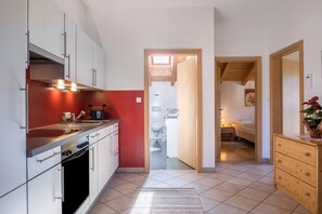 A compact kitchen with white cabinets, a black oven, and a red accent wall. A wooden dresser and a small table with a vase are visible in the foreground.