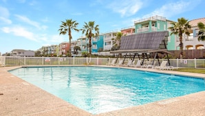 A swimming pool with lounge chairs and a shaded area, surrounded by colorful buildings and palm trees.