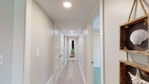 A hallway with white walls, wooden flooring, and a ceiling light. There are doors on both sides, and a decorative shelf with shells on the right.