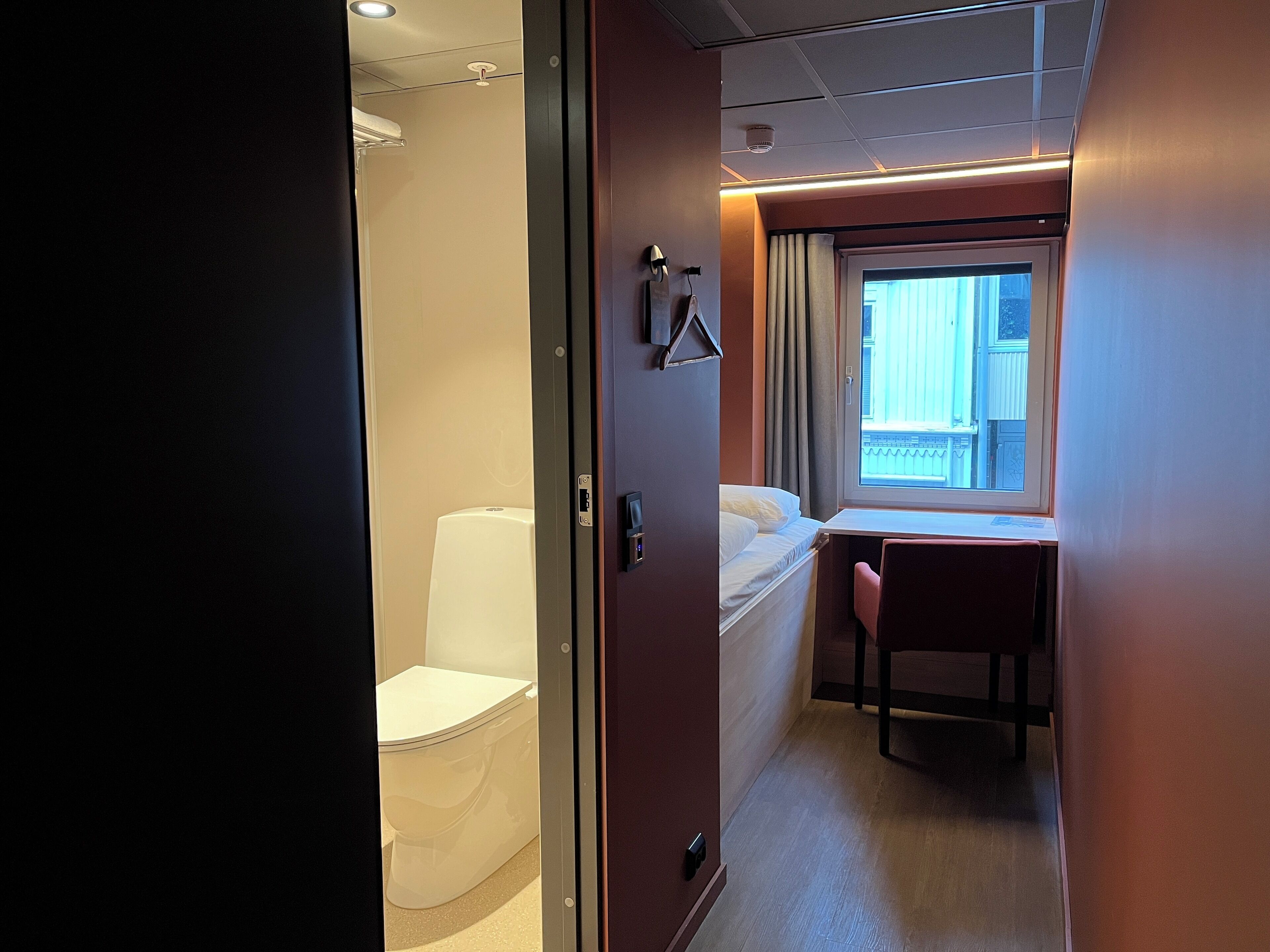 A narrow hallway with a bed, a desk, and a toilet.
