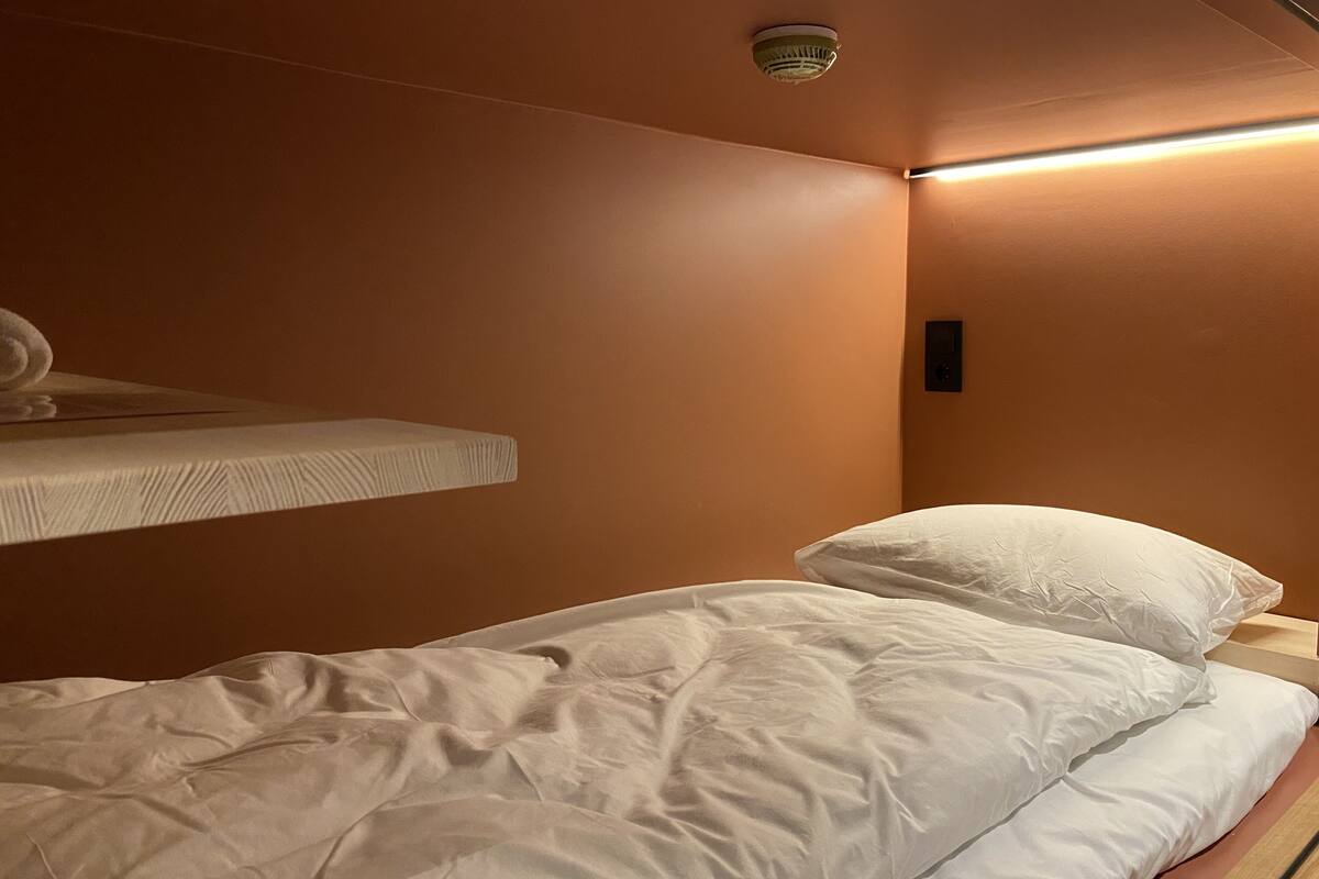 Uma cama de solteiro com roupa de cama branca em quarto com paredes marrons e luminária no teto.