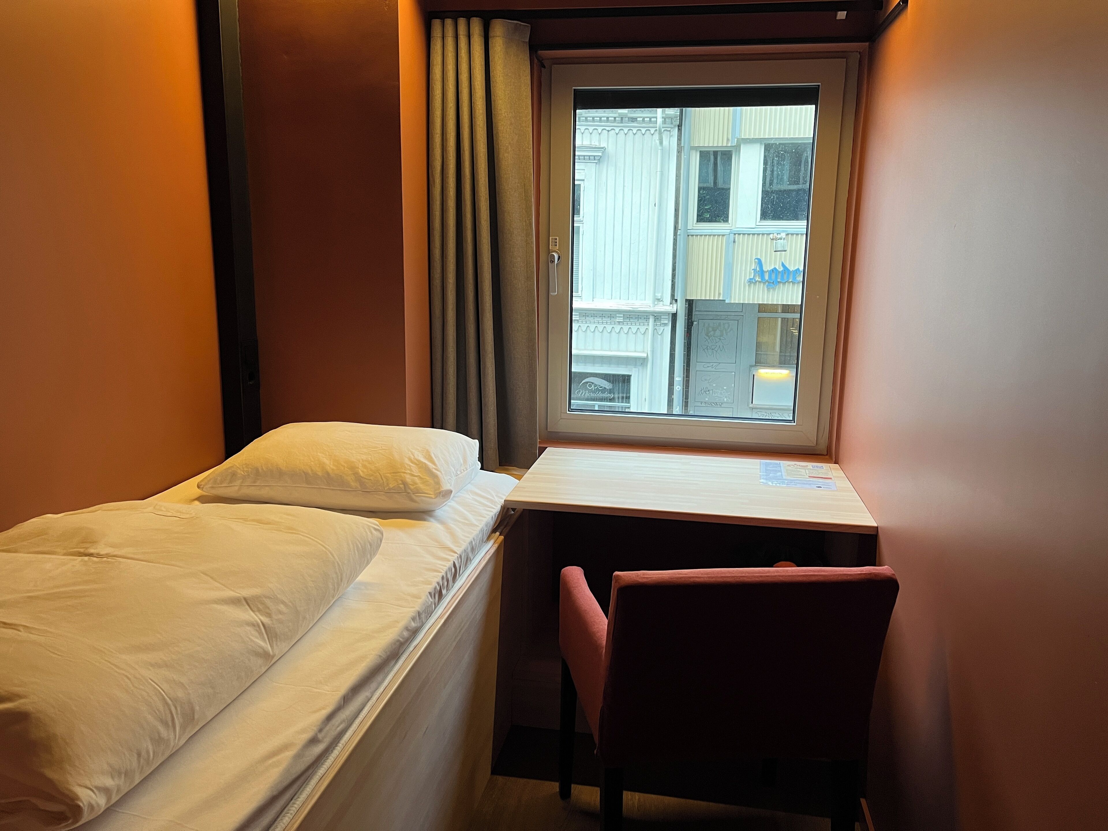 Habitación de hotel pequeña con una cama individual, un escritorio y una silla. Se ve una ventana con vistas a edificios.