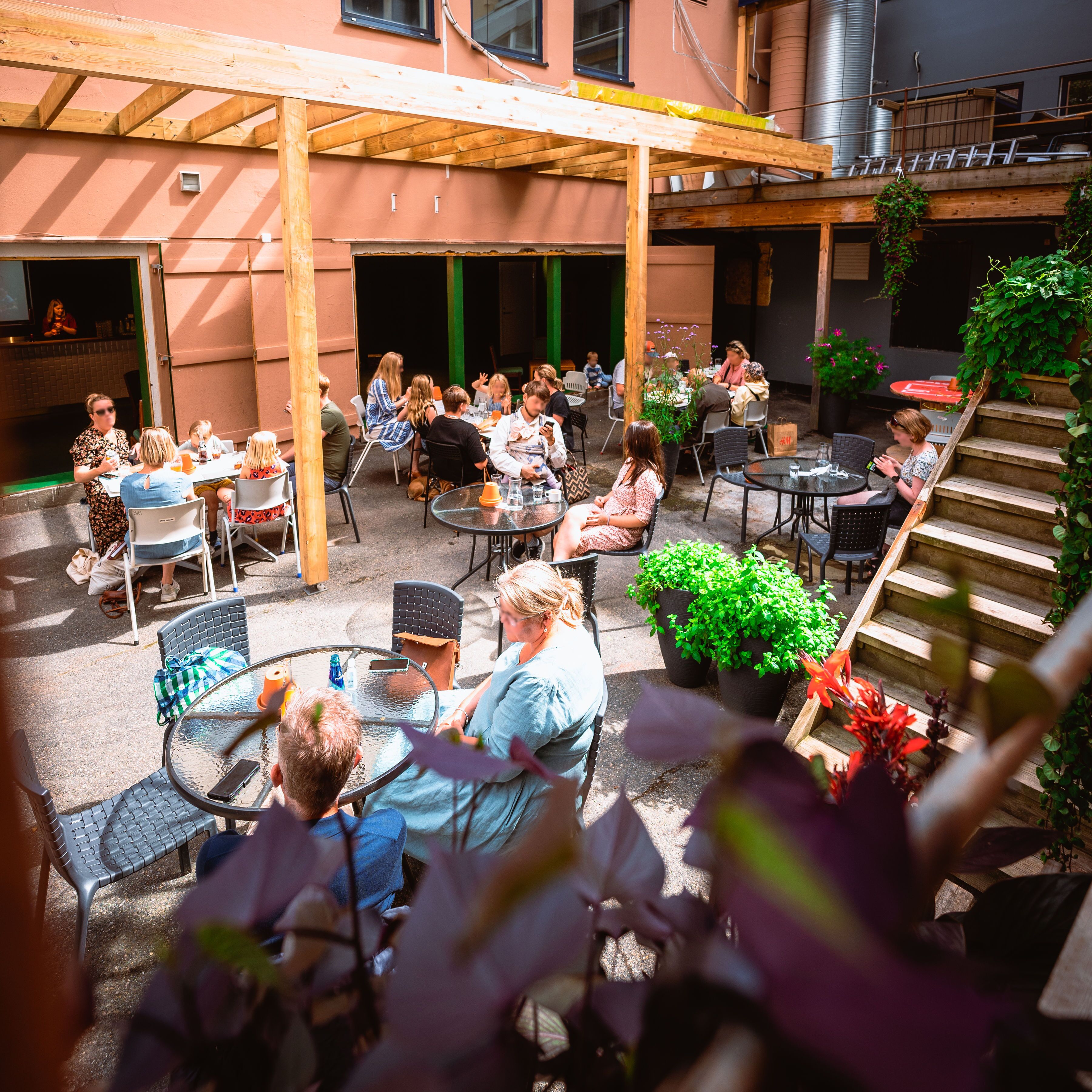 An outdoor patio area with multiple tables and chairs, surrounded by plants and a wooden pergola.
