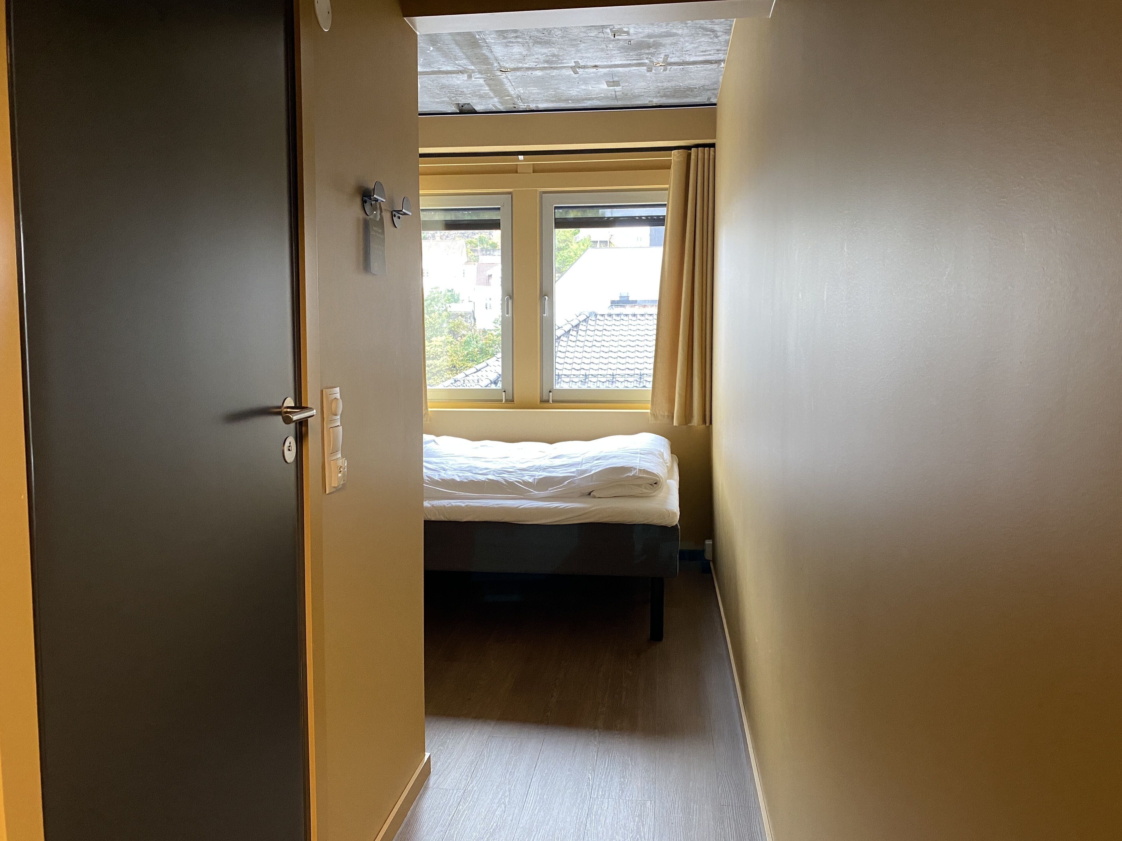 A narrow hallway leading to a bedroom with a bed, a window with curtains, and a door.