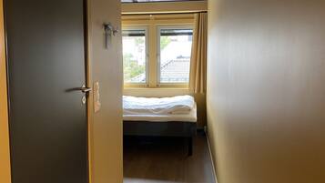 A narrow hallway leading to a bedroom with a bed, a window with curtains, and a door.