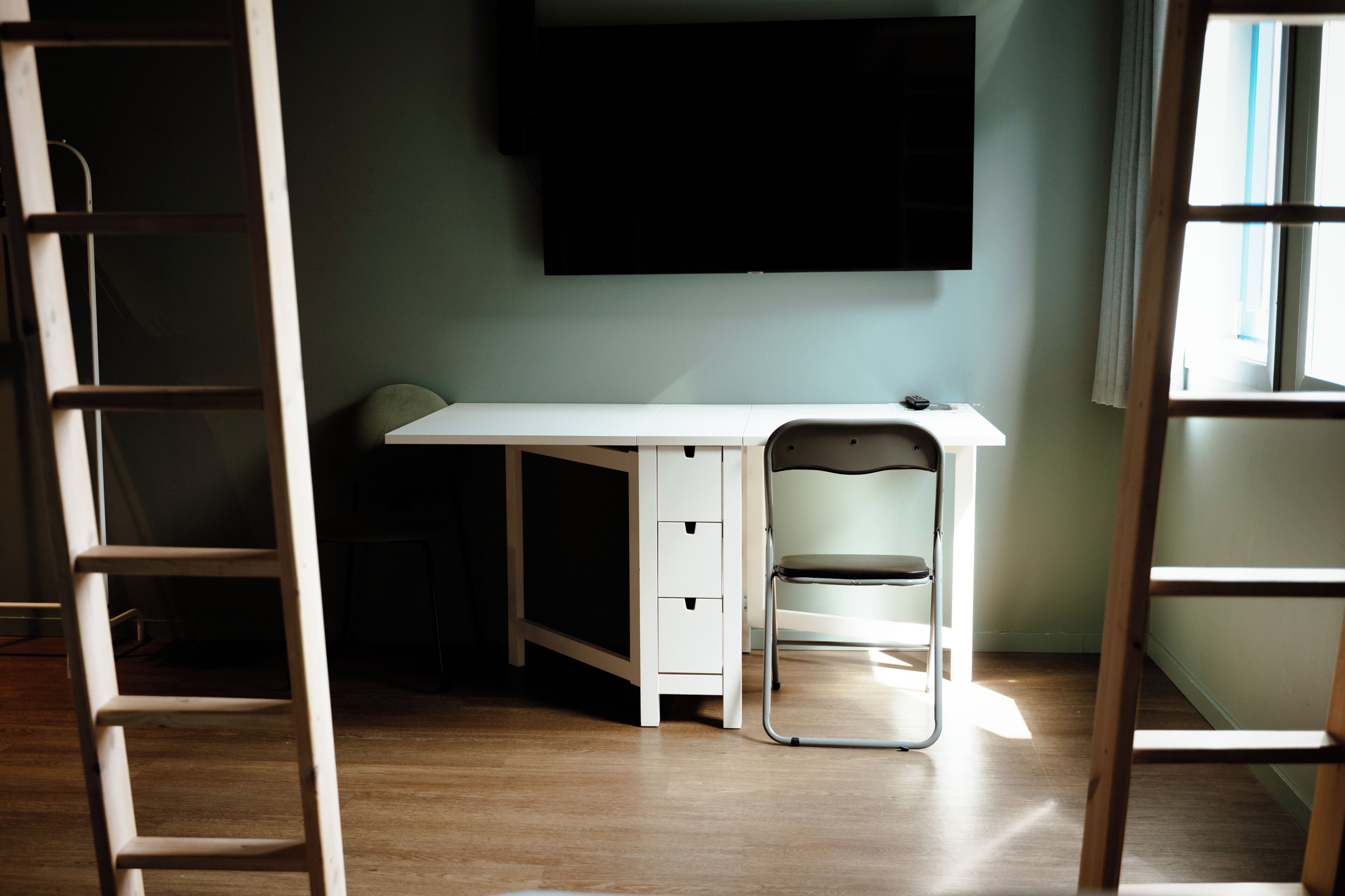 Une pièce avec un bureau blanc, une chaise, une échelle et un téléviseur fixé au mur.