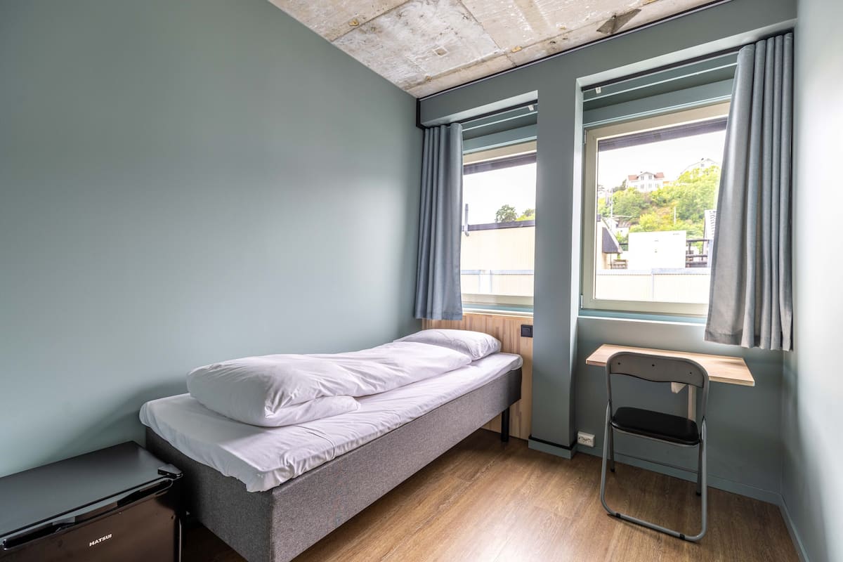 Quarto pequeno com uma cama de solteiro, piso de madeira, escrivaninha e janela com cortinas.