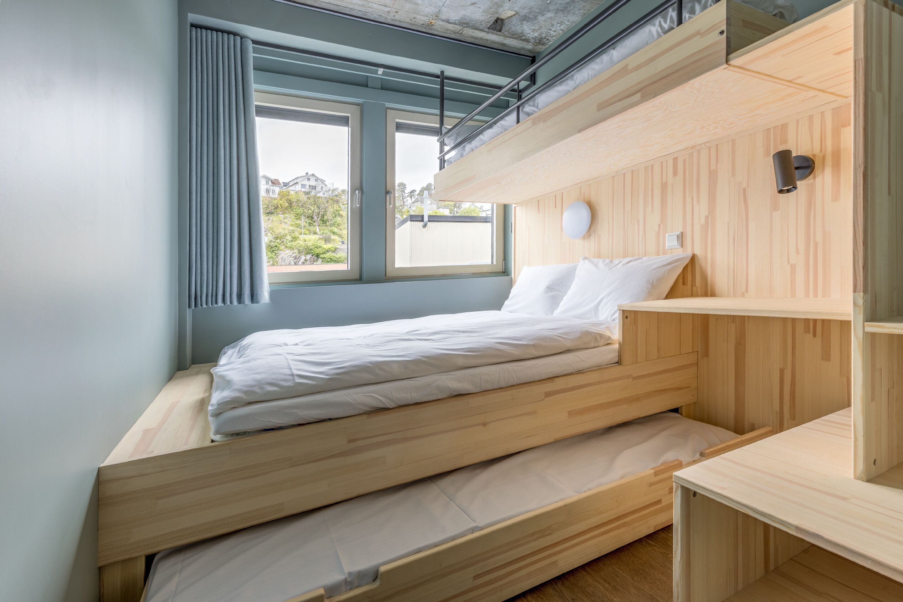 A compact, modern room with a bunk bed, a small desk, and a window with a view of buildings and greenery.