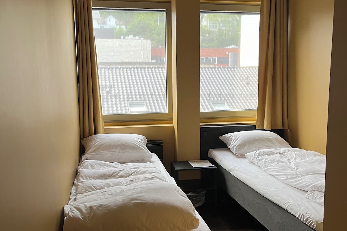 Quarto pequeno e compacto com duas camas de solteiro, uma mesa de cabeceira e janela com cortinas.