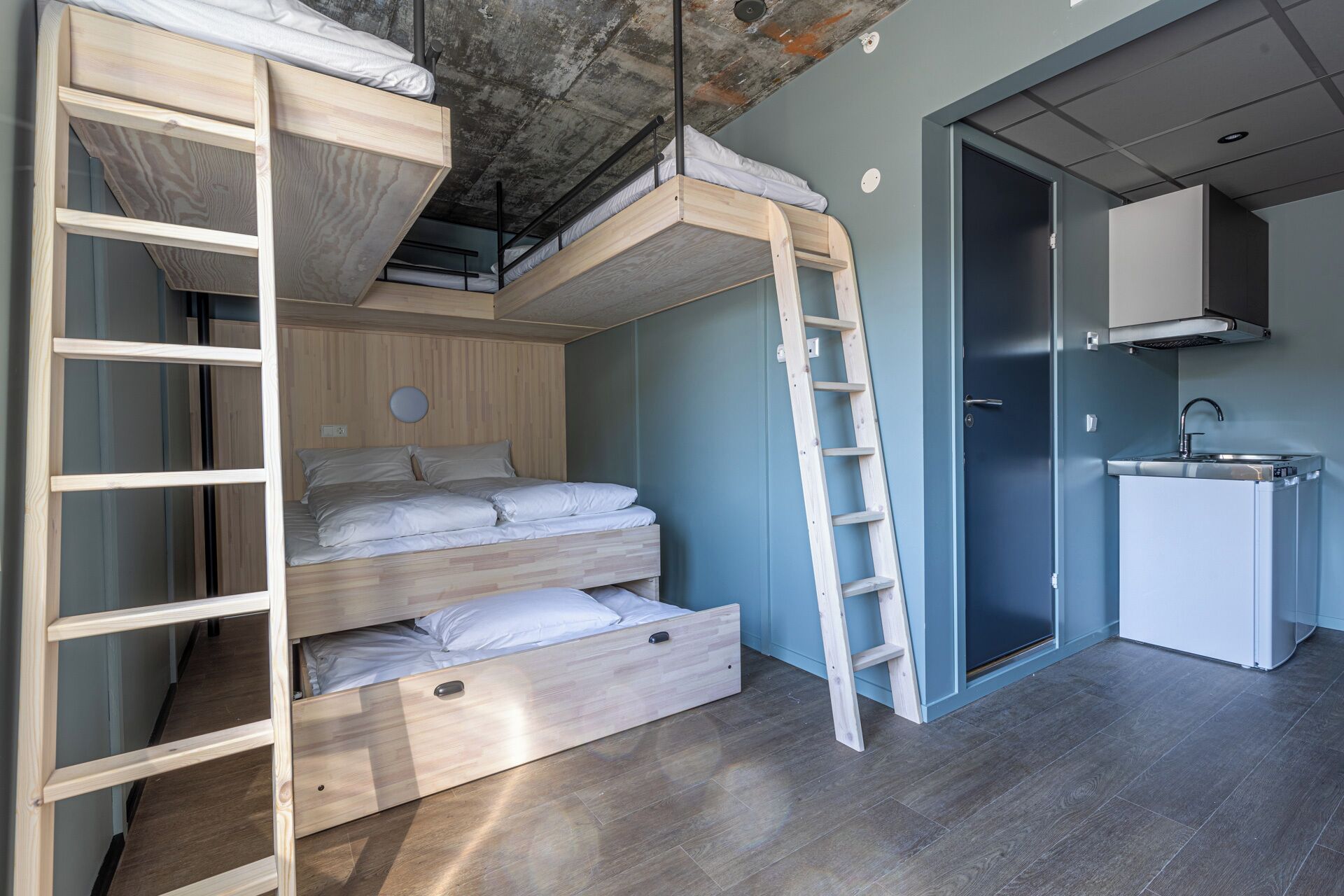 Une pièce compacte comprenant un lit superposé, une échelle, une petite kitchenette et une porte donnant sur une autre pièce.