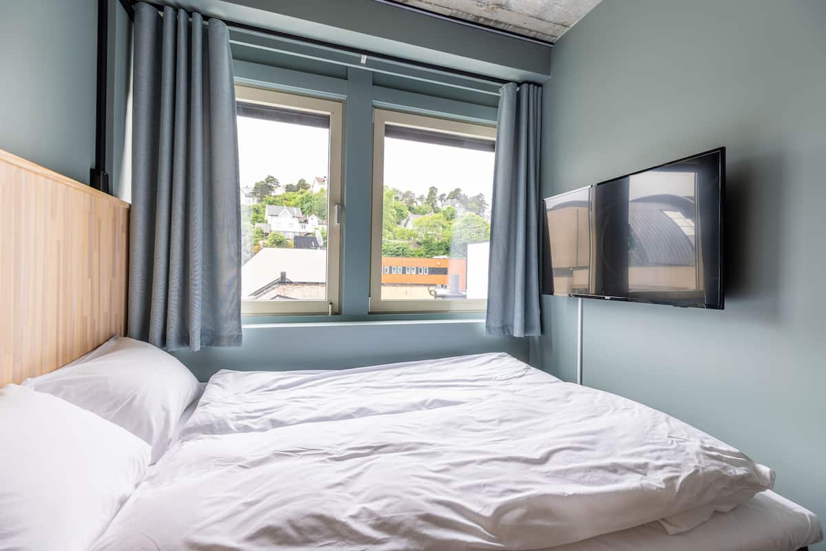 Quarto com cama, janela com cortinas e televisão presa na parede.