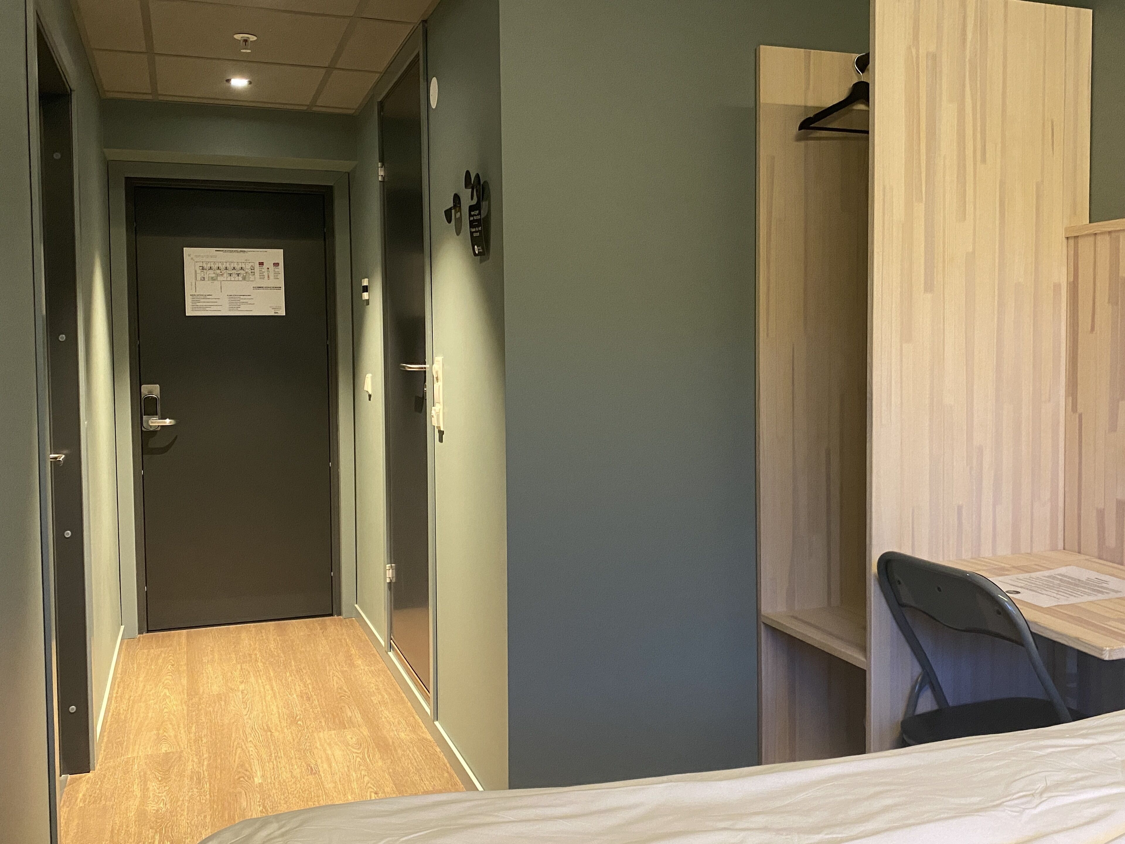 A hotel room with a bed, a desk, and a chair. There is a door at the end of the hallway.
