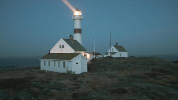 Un faro con un edificio blanco cercano, ambos ubicados en terreno rocoso con vista al mar.