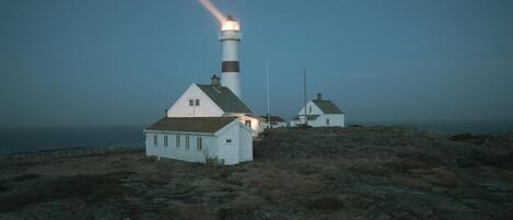 A lighthouse with a white building nearby, both situated on a rocky terrain with a view of the sea.