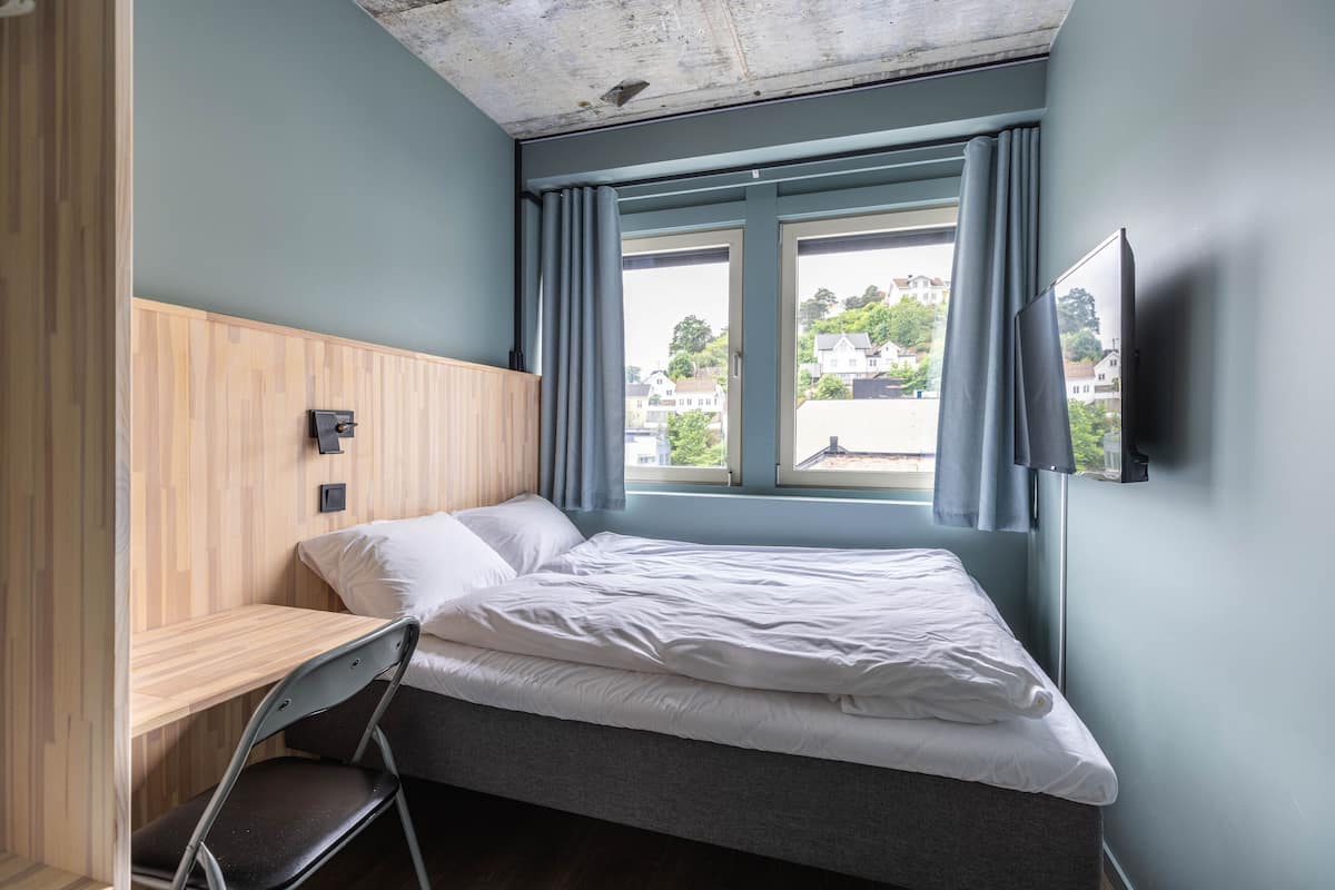 Uma cama de solteiro com cabeceira de madeira, uma escrivaninha com cadeira, uma televisão de tela plana na parede e uma janela com cortinas.