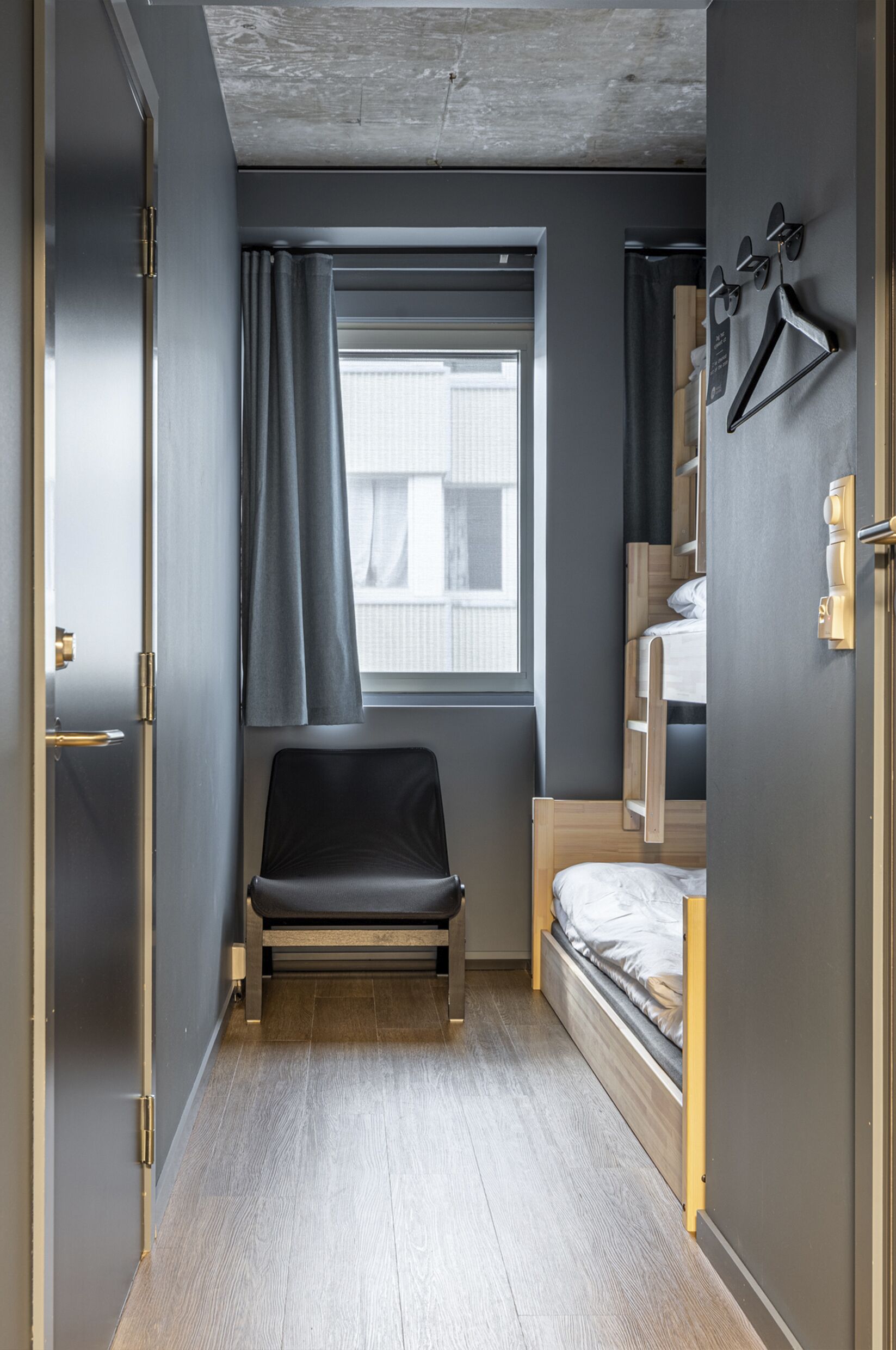 Habitació petita amb un llit individual, una cadira, una finestra amb cortines i una perxa per a abrics.