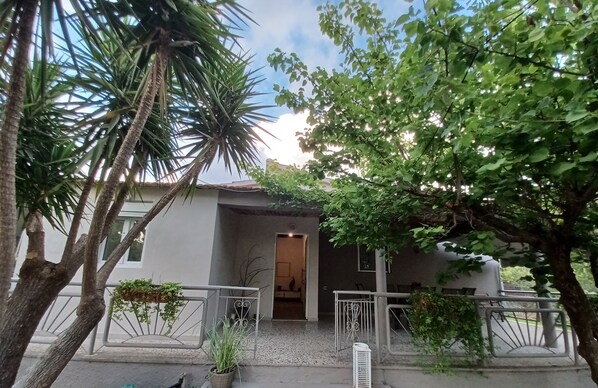 A modern building with a covered entrance, surrounded by trees and a small garden area with potted plants.