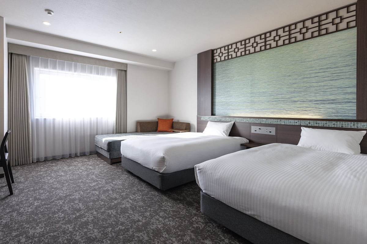 Habitación de hotel con dos camas, un ventanal con cortinas, un televisor montado en la pared y una alfombra estampada.