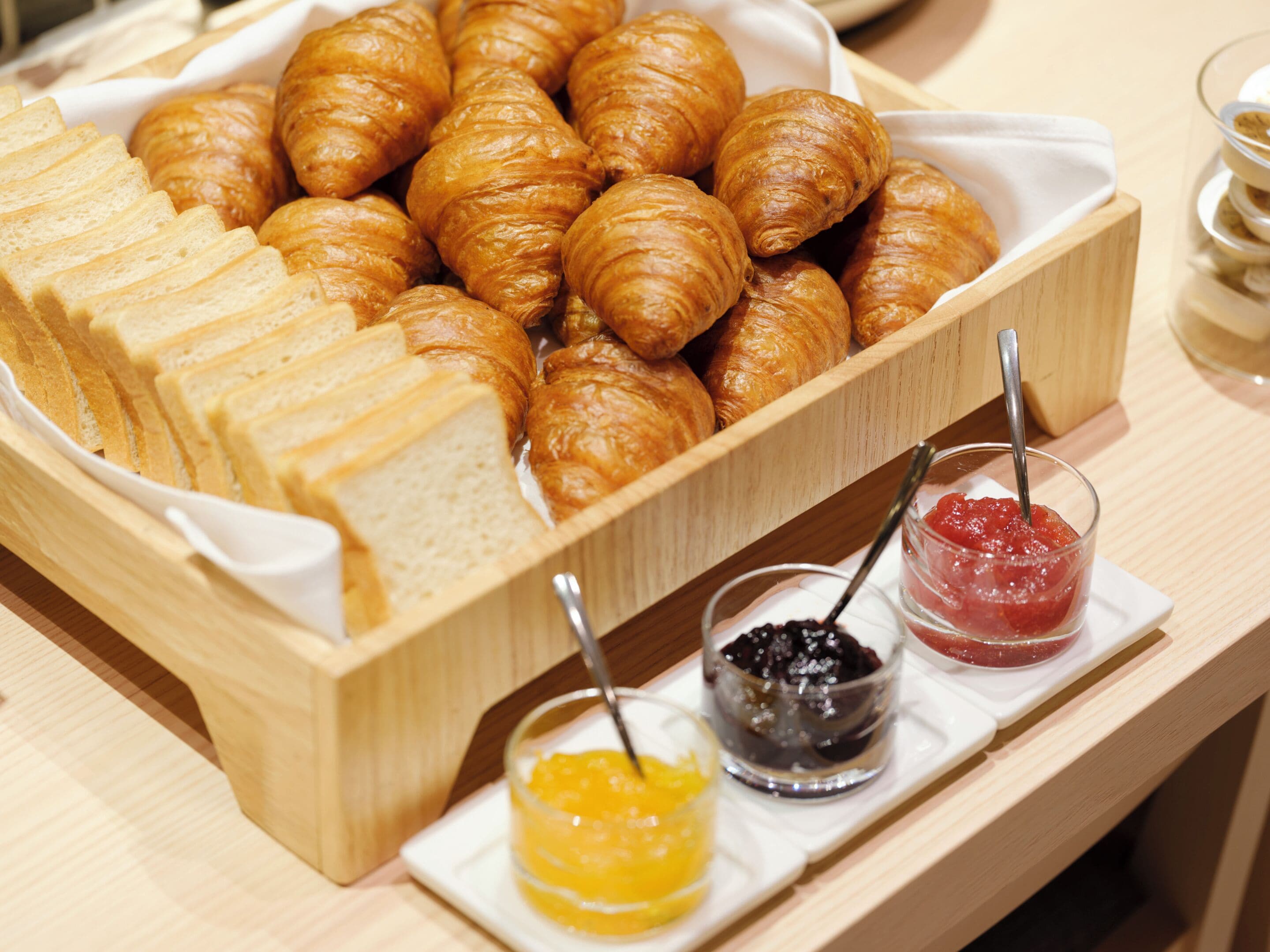 A wooden serving tray with croissants and sliced bread, accompanied by two small bowls of jam.