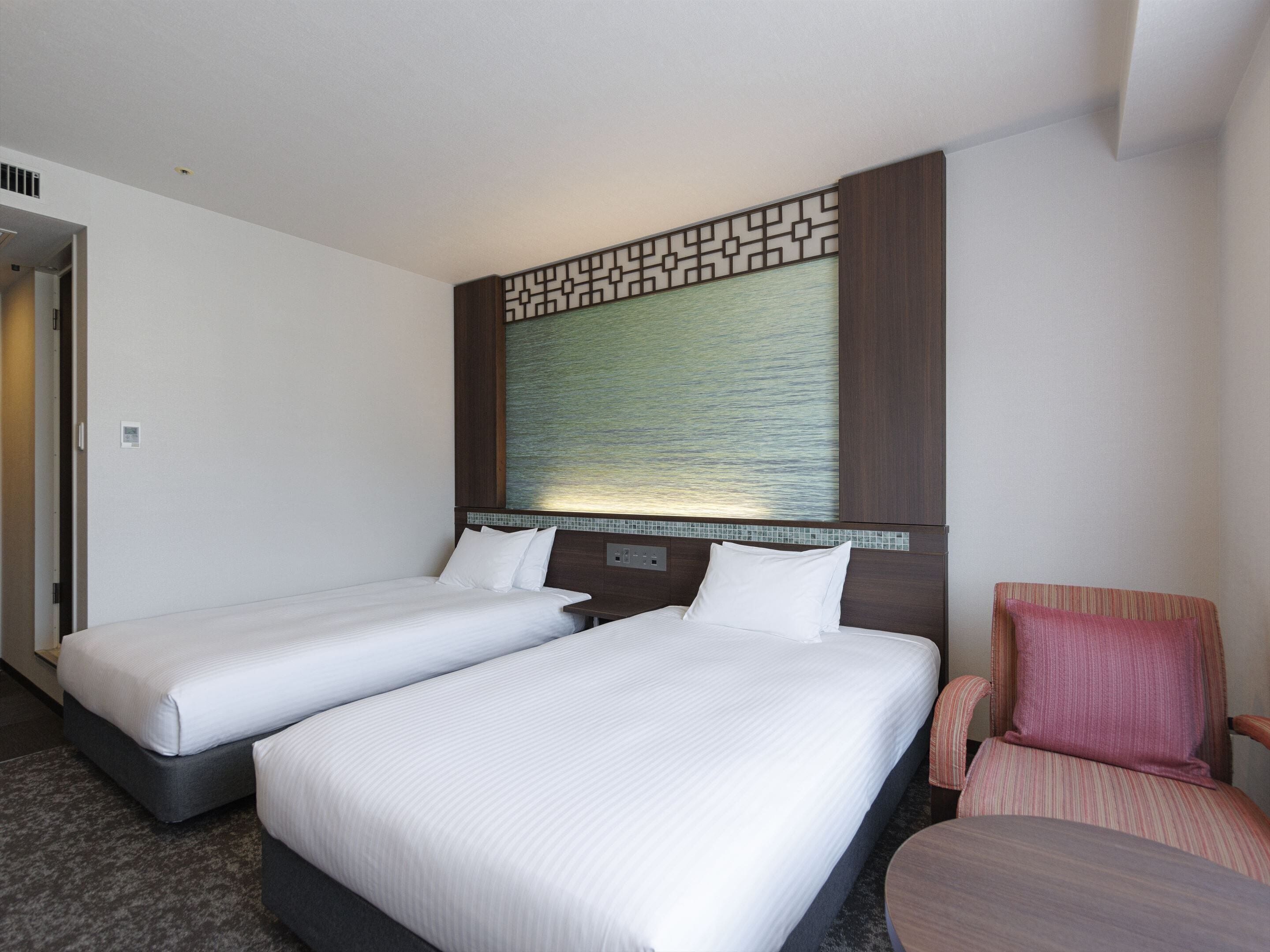 A hotel room with two single beds, a small seating area with a chair, and a decorative wall panel above the beds.