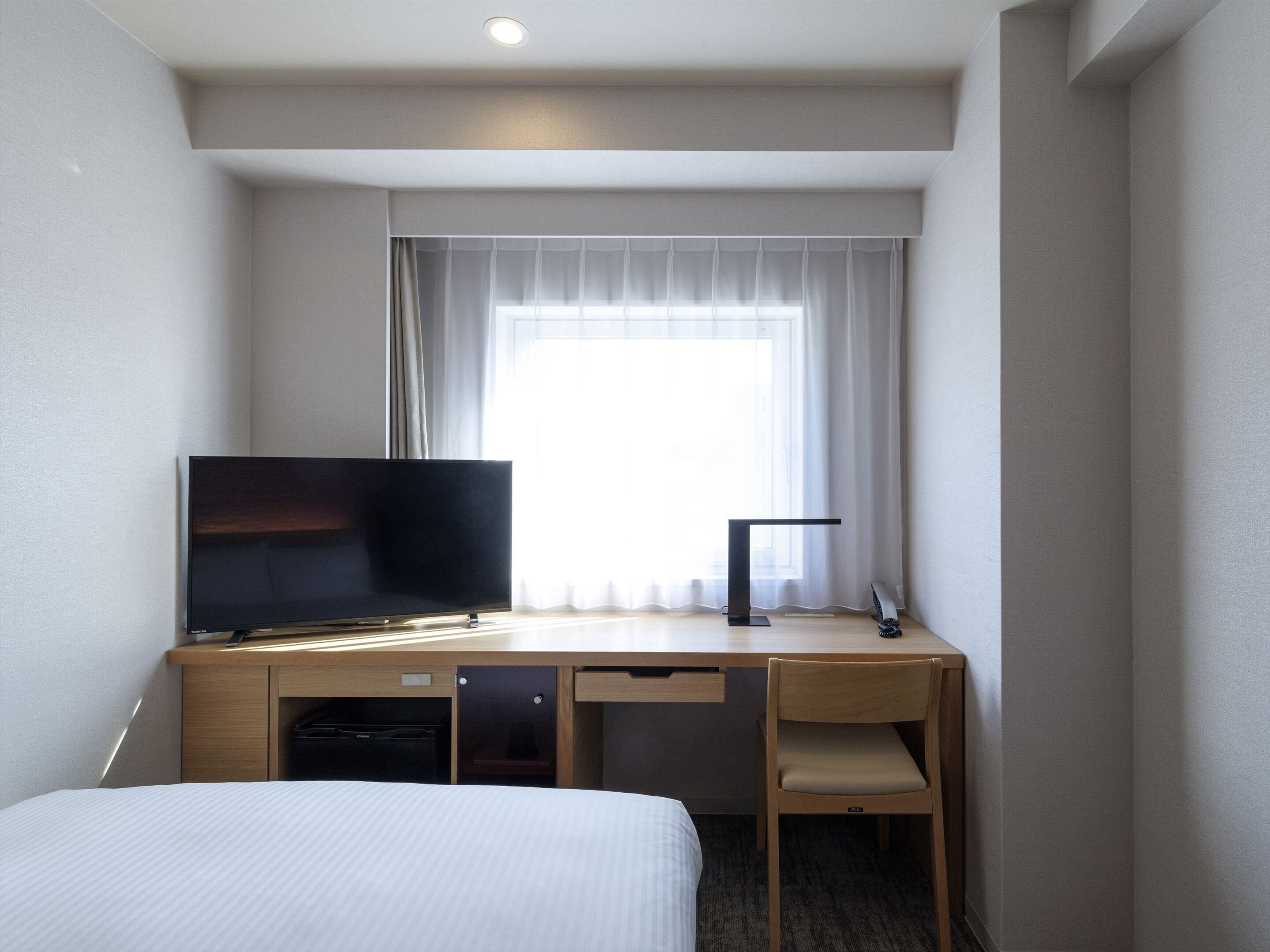 A hotel room with a bed, a desk with a TV, a chair, and a window with curtains.