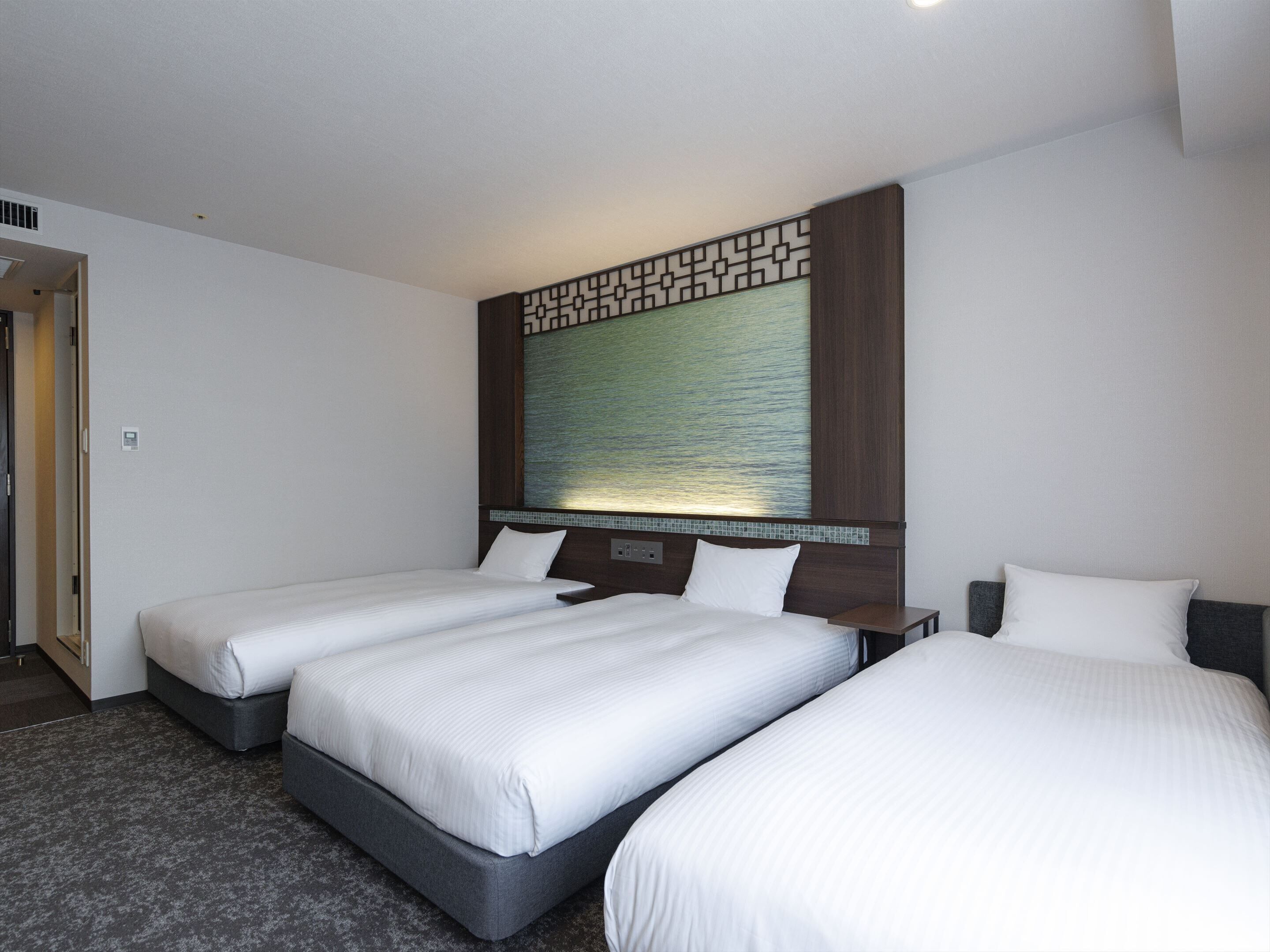 A hotel room with two single beds and one double bed, a large window with a view, and a decorative wall panel.