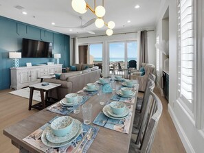 A modern living room with a dining area set for a meal, featuring a large table with blue tableware, a flat-screen TV, and a view of the ocean. - Hook, Wine, and Sinker Luxurious 4 bedroom Townhome! (Navarre Beach)