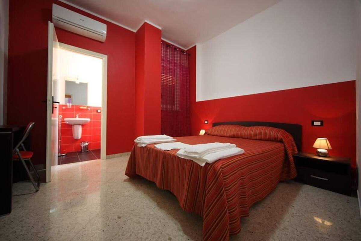 Quarto de hotel com cama, mesa de cabeceira, luminária, cadeira e banheiro visível por uma porta aberta.