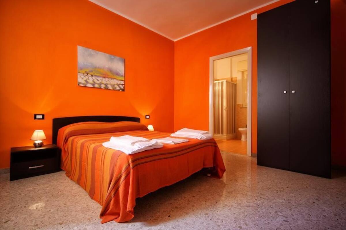 Quarto de hotel com parede laranja, cama com roupa de cama listrada, mesa de cabeceira com abajur e um quadro acima da cama.